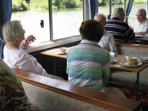 Fünf Ausflugsteilnehmer des Nachmittagskreises sitzen im Boot an Tischen bei Kaffee und Kuchen; durch die Glasscheibe fällt der Blick auf den Neckar und das grüne Ufer